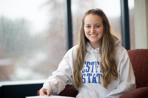 Class of 2024 President, Kaitlyn Egan, sitting on a red chair by a window. She has long blond hair. She wears a gray sweatshirt that says "Westfield State" on it and smiles.