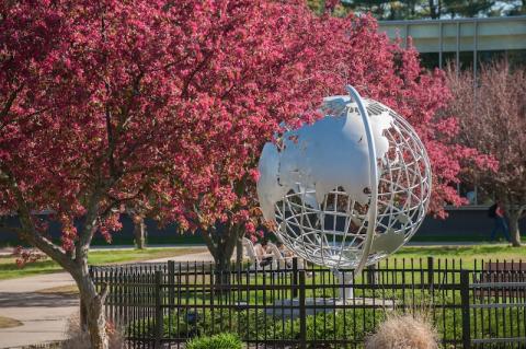 A photo of the silver, campus globe. Trees bud beside it, sprouting pink flowers. Green grass is behind the globe, and a black wrought-iron fence surrounds it.