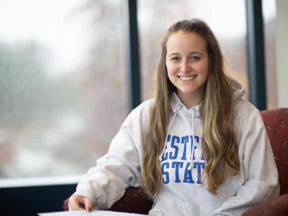 Class of 2024 President, Kaitlyn Egan, sitting on a red chair by a window. She has long blond hair. She wears a gray sweatshirt that says "Westfield State" on it and smiles.