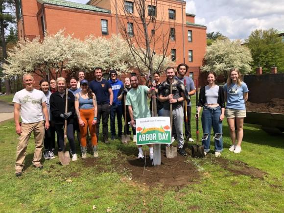 This year's Arbor Day. Students and staff from the biology department gather around a newly planted tree. A white and green sign saying "Arbor Day" is also planted in front of the tree.