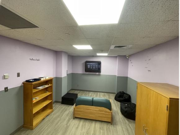 A new pediatric waiting room at the MiraVista Behavioral Health Center in Holyoke, MA, as part of the Psychiatric Nursing class. The walls are half-white and half-green, with a wood book shelf, cabinet, and several bean bags dotting the floor in front of a TV.