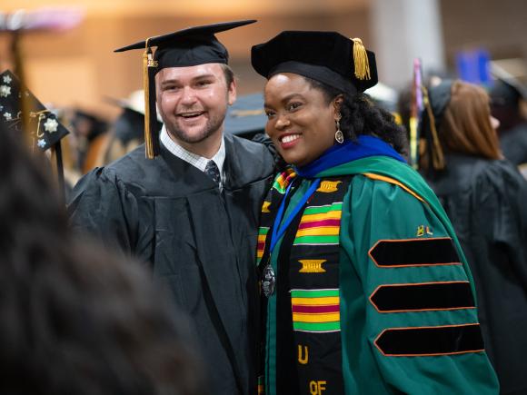 Education faculty member and student wearing caps and gowns at commencement smiling.