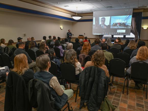 Faculty member speaking at a podium during a campus lecture with a group of attendees seated in chairs.