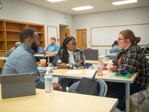 Three students engaged in discussion while seated together in a classroom.