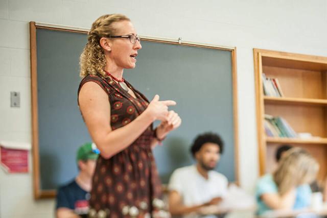 A teacher stands amongst seated students in a classroom providing instruction.