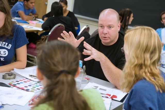 A math professor sits at a table with a group of students to discuss a class assignment.