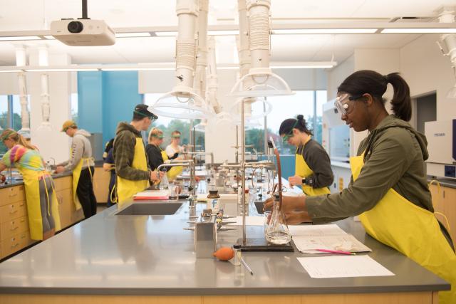 Chemistry lab with students learning hands on wearing lab aprons and eye protection.