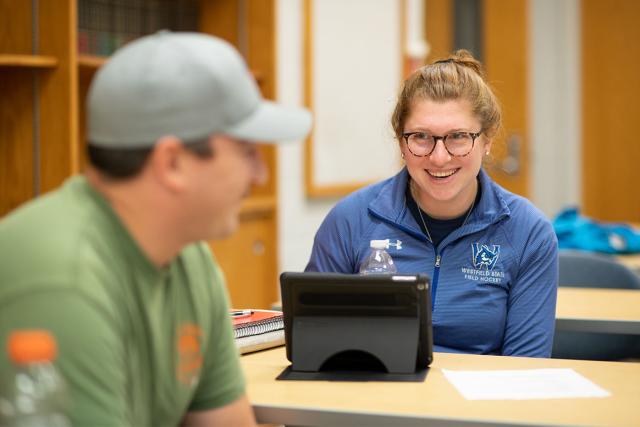 Two students smile and laugh while talking in a classroom.