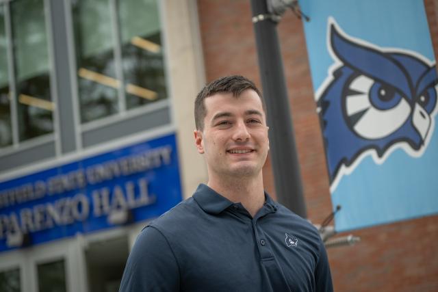 Student smiling outside of Parenzo Hall with an owl flag behind him.