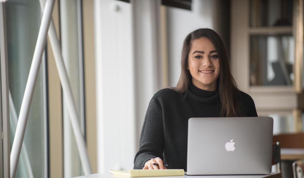 female student smiling at the camera as they work on their laptop in the Ely Campus Center