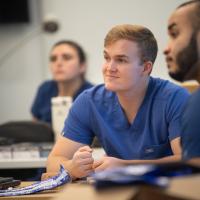 Three students in classroom wearing blue scrubs with WSU logos listening to instructor.