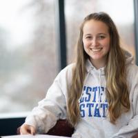 Class of 2024 President, Kaitlyn Egan, sitting on a red chair by a window. She has long blond hair. She wears a gray sweatshirt that says "Westfield State" on it and smiles.