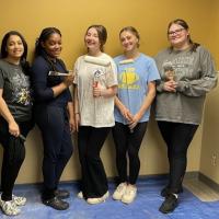 The five students who created a new pediatric waiting room at the MiraVista Behavioral Health Center in Holyoke, MA. Four young women pose in front of a yellow wall, and they're holding paint brushes to show some of the work they've done to create the room.