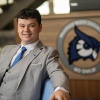 Westfield State student in a gray suit smiles while seated in front of a university podium featuring the Westfield State owl logo.
