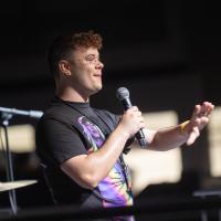Student speaks into a microphone onstage during Westfield State’s Spring Weekend, gesturing to the audience while wearing colorful face gems.